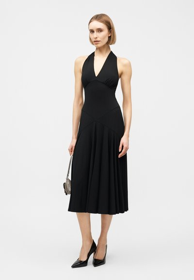 Femme portant une robe noire sans manches à col licou avec une jupe évasée, des escarpins noirs pointus et tenant un petit sac gris, debout devant un fond blanc.