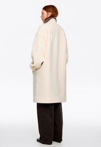 Persona con cabello rojo que lleva un abrigo largo de lana color crema con cuello alto y pantalones negros de pierna ancha, de pie y ligeramente girada hacia la izquierda.