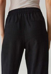 Schwarze Hosen aus strukturiertem Stoff, mit einem elastischen Bund und zwei Gesäßtaschen, von hinten betrachtet.