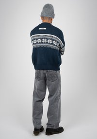 Homme portant un bonnet en maille gris, un pull bleu marine avec un motif nordique blanc, un jean gris ample et des chaussures noires, debout de dos sur un fond gris.