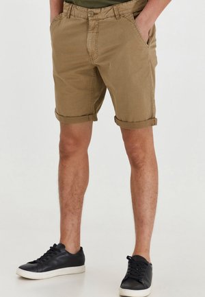 Hombre vestido con pantalones cortos de caqui remangados y zapatillas negras, de pie con las manos en los bolsillos sobre un fondo blanco liso.