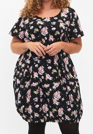 Kvinde iført en sort kjole med korte ærmer med pink, hvide og gule blomsterprint, hænderne foldet foran og krøllet hår synligt.