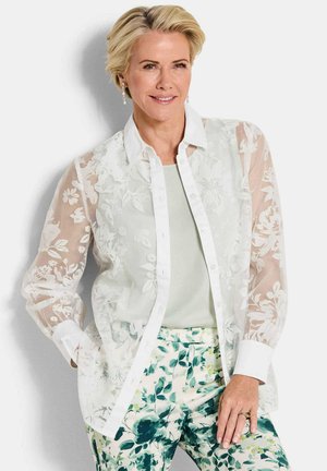 Femme souriante aux cheveux blonds courts portant un chemisier blanc transparent brodé de fleurs sur un haut clair et un pantalon à motifs floraux verts.