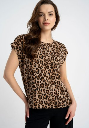 Kobieta o długich brązowych włosach, ubrana w krótki top w panterkę i czarne spodnie, pozująca z jedną ręką na biodrze.