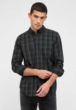 Młody mężczyzna podwijający rękawy czarno-zielonej koszuli w kratę z guzikami, stojący na tle jednorodnego jasnoszarego tła.