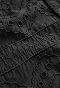 Sort stof med indviklet broderi, bølgete kanter og cirkulære udskæringer, der viser mærkenavnet "ALLSAINTS" i tekstureret syning.