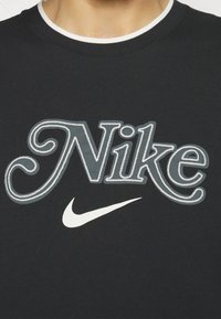 Svart bomullströja med "Nike"-logotypen i en dekorativ, utmattad stil och en vit Nike swoosh-logotyp. Rund ribbad halsringning.