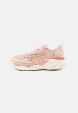 Zapatilla deportiva de color rosa claro con parte superior de tejido de punto, suela blanca gruesa, nombre de la marca en relieve en el lateral y talón esculpido para amortiguación y soporte.