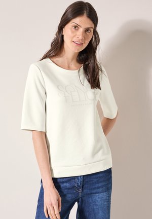 Sweatshirt beige clair à manches courtes, décolleté en U, et texte en relief "DÉCOUVRIR & APPRECIER." Associé à un jean bleu foncé. Texture douce.