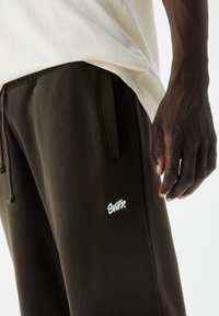 Mörkbruna träningsoveraller i mjukt material, med en vit broderad logotyp och justerbar dragsko vid midjan.