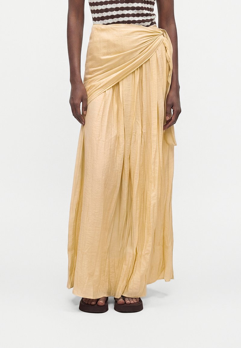 Femme portant une jupe longue plissée jaune clair avec un détail de nœud froncé et des sandales marron foncé à bout carré.