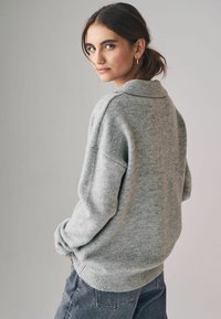 Maglione grigio oversized a maglia con un ampio colletto e polsini a costine. Indossato sopra jeans scuri, crea un look casual a strati.