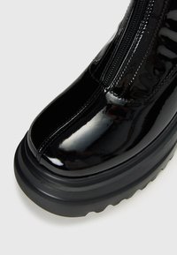 Glossy black patent leather boot with thick ridged sole and visible front zipper on a plain light background.
