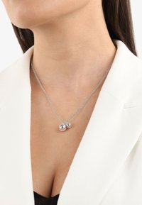 Collana d'argento con due pendenti rotondi, uno più grande e riflettente, appesi a una catena delicata sopra un blazer bianco.