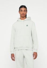 Fiatal férfi, aki fonott hajat visel, világosszürke Adidas kapucnis pulóverben és hozzá illő tréningnadrágban áll a fehér háttér előtt.
