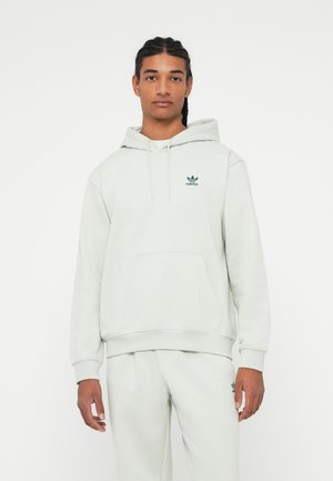 Junger Mann mit geflochtenem Haar, der einen hellgrauen Adidas-Hoodie und passende Jogginghosen trägt, steht vor einem weißen Hintergrund.
