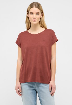 Junge Frau mit langen blonden Haaren, die ein rostfarbenes kurzärmeliges T-Shirt und hellblaue Jeans trägt, steht vor einem weißen Hintergrund.