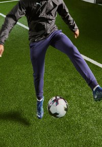 Černá sportovní bunda, tmavě modré tepláky a modré fotbalové kopačky na travnatém hřišti, zaměřeno na nohu ovládající černobílý fotbalový míč.