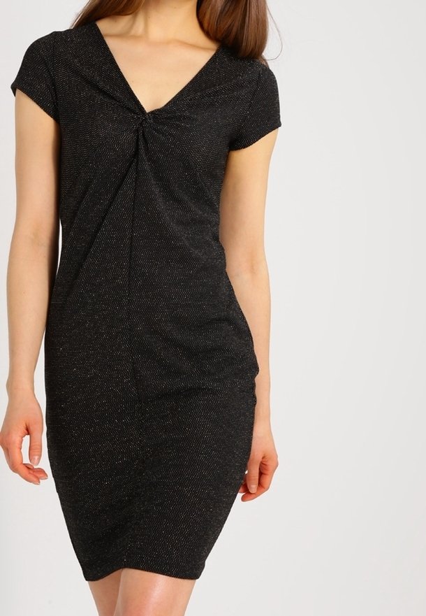 Femme portant une robe noire ajustée à manches courtes avec un détail de nœud torsadé sur la poitrine, sur un fond clair uni.