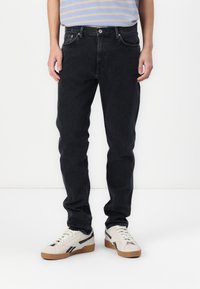 Jean en denim noir avec une coupe droite, dotés de cinq poches et de coutures contrastées. Portés avec des baskets blanches et une chemise rayée.
