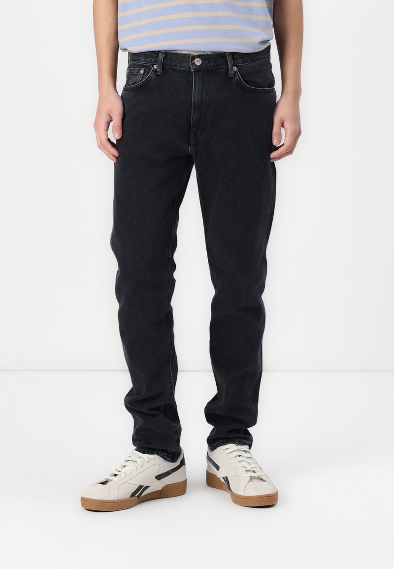 Jean en denim noir avec une coupe droite, dotés de cinq poches et de coutures contrastées. Portés avec des baskets blanches et une chemise rayée.