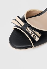 Czarny skórzany but z otwartym palcem, ozdobiony beżową kokardą i logo. Gładka faktura z detalami w kontrastującym kolorze.