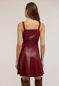Donna con lunghi capelli castani, vestita con un abito in pelle borgogna senza maniche e stivali marroni al ginocchio, in piedi di spalle su uno sfondo neutro.