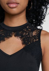 Haut noir sans manches avec un corsage texturé, des détails en dentelle autour de l'encolure et une ouverture en forme de goutte d'eau. Finition en tissu lisse.