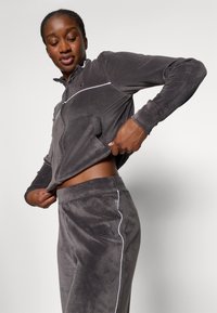 Giacca in velluto grigio con zip e piping bianco, abbinata a pantaloni a vita alta coordinati. Presenta tasche e una vestibilità rilassata, con una texture liscia.