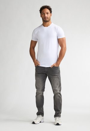 T-shirt blanc à manches courtes avec un col rond, associé à un jean gris et des baskets blanches, debout sur un fond uni.