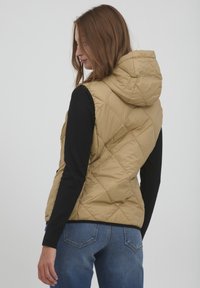 Khaki-Steppweste mit Kapuze, diagonal gequiltet, mit Stehkragen und seitlichen Taschen. Getragen über einem schwarzen Langarmshirt.