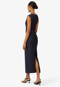 Marineblaues, figurbetontes Kleid mit kurzen Ärmeln und Reißverschluss hinten, das einen seitlichen Schlitz und ein elegantes Design aufweist. Getragen mit schwarzen, hohen Sandalen.