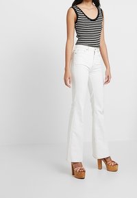 Schwarz-weißer gestreifter Tanktop, weiße Schlagjeans und braune Plateau-Pumps mit abgerundeter Spitze und Riemchendetails. Weiche Stofftexturen.