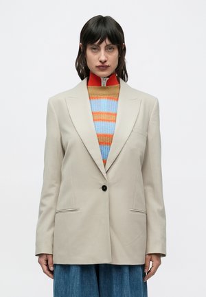 Beiger, übergroßer Blazer mit einem schwarzen Knopf, gekerbtem Revers, Vorderentaschen, getragen über einem mehrfarbig gestreiften Pullover.