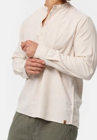 Mężczyzna w beżowej koszuli z długim rękawem i kołnierzykiem mandarynkowym, zapinanej pod szyją, poprawia mankiety dłońmi, w połączeniu z teksturowanymi oliwkowymi spodniami.
