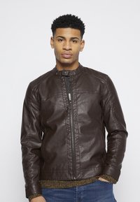 Chaqueta de cuero marrón oscuro con cuello alto y cierre de cremallera, que presenta detalles de costura sutiles y una textura suave.