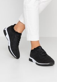 Schwarze Sportschuhe mit einem strukturierten Strickobermaterial, weißer Sohle und geripptem Kragen. Mit schwarzen Schnürsenkeln und einem eleganten, modernen Design.