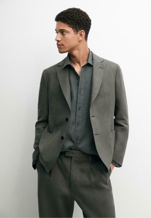 Junger Mann trägt einen grauen Anzug mit passendem Hemd, steht mit Händen in den Taschen und schaut nach links vor einem einfarbigen weißen Hintergrund.