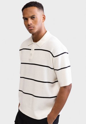 Mand står med hænderne i lommerne, iført en hvid kortærmet poloshirt med sorte vandrette striber, vendt fremad mod en ensfarvet baggrund.