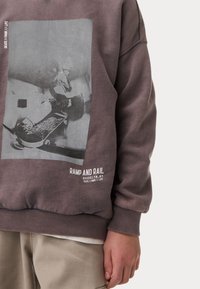 Felpa in tessuto viola tenue, con una grande grafica grigia di un skateboarder. Il testo in basso recita "RAMP AND RAIL" in bianco.