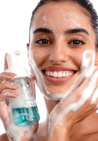 Schuimende reiniger in een fles met een witte pomp, helder plastic, turquoise vloeistof, omringd door gezichtsfoam op de huid en handen, model glimlachend.