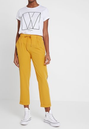 Frau trägt ein weißes, hochgekrempeltes T-Shirt mit Grafik, senfgelbe Hose mit Kordelzug und weiße High-Top-Sneaker und steht vor einem weißen Hintergrund.