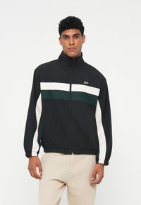 Jachetă neagră tip windbreaker cu guler înalt, având dungi orizontale albe și verzi. Mansete și tiv elastic. Logo-ul Lacoste pe piept.