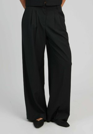 Personne portant un pantalon large noir et ample et des ballerines noires à bout pointu, se tenant contre un fond clair uni.