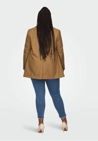 Persoon met lang gevlochten haar, gekleed in een bruine blazer, blauwe strakke jeans en beige hoge hakken, staand met de rug naar de camera op een witte achtergrond.