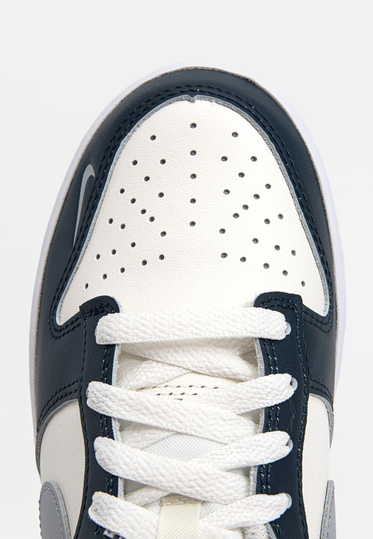 Sneaker bianco e blu marina con tomaia in pelle perforata, lacci bianchi piatti e un accento grigio. Presenta un logo cucito e una suola testurizzata.