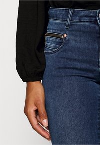 Skinny blue denim jeans with a zippered pocket detail, high waistband, and slight fade, paired with a black textured top and a hand resting on the thigh.