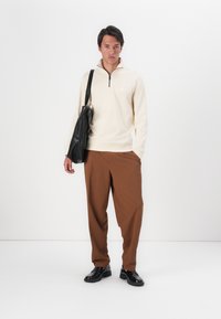 Cream zip-up sweater with a stand collar, brown pleated trousers, and black leather bag. Model wears black shoes, standing against a white background.