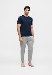 Homme debout pieds nus, portant un t-shirt bleu marine à manches courtes et un pantalon de survêtement gris clair, les mains dans les poches, sur un fond uni.
