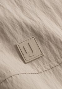Tessuto beige testurizzato con un patch in gomma rettangolare e lettere in rilievo "UJ" e dettagli di cucitura visibili.